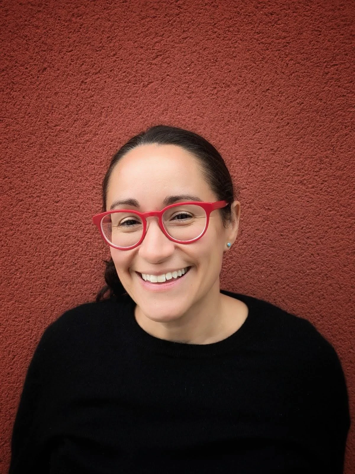 Catherine Hulshof, smiling, in red glasses against a red wall.
